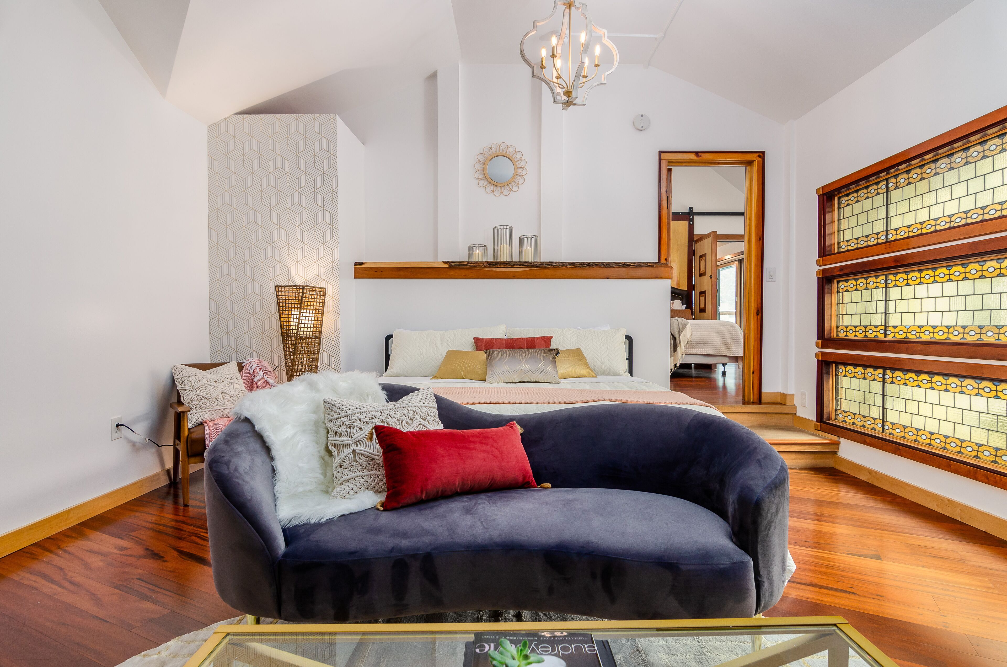 A modern living room with a dark blue sofa, a white fluffy throw, and a red pillow. There is a wooden coffee table with a magazine. A staircase leads to another room with a wooden railing and a large mirror.