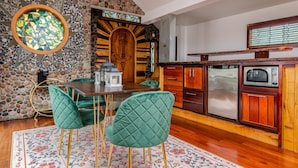 A dining area with a wooden table and green upholstered chairs, a stone wall, and a kitchen with wooden cabinets and appliances. - New Park Event Venue & Suites (Ithaca)
