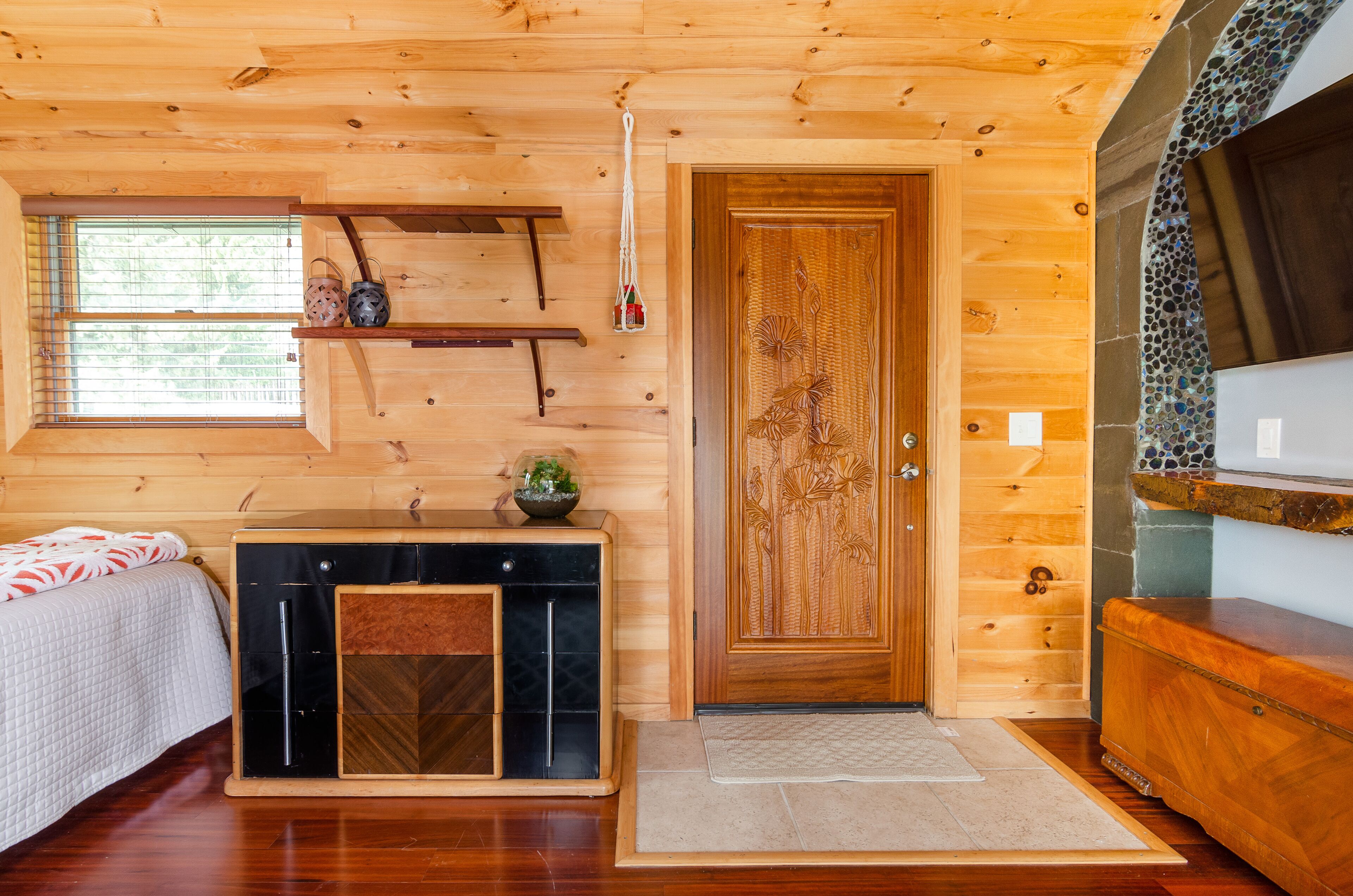 A wooden cabin interior with a bed, a console table, a door, and a window with blinds.