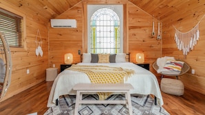 A cozy wooden cabin bedroom with a large bed, a bench, a window with a stained glass design, and a hanging macrame decoration. - New Park Event Venue & Suites (Ithaca)