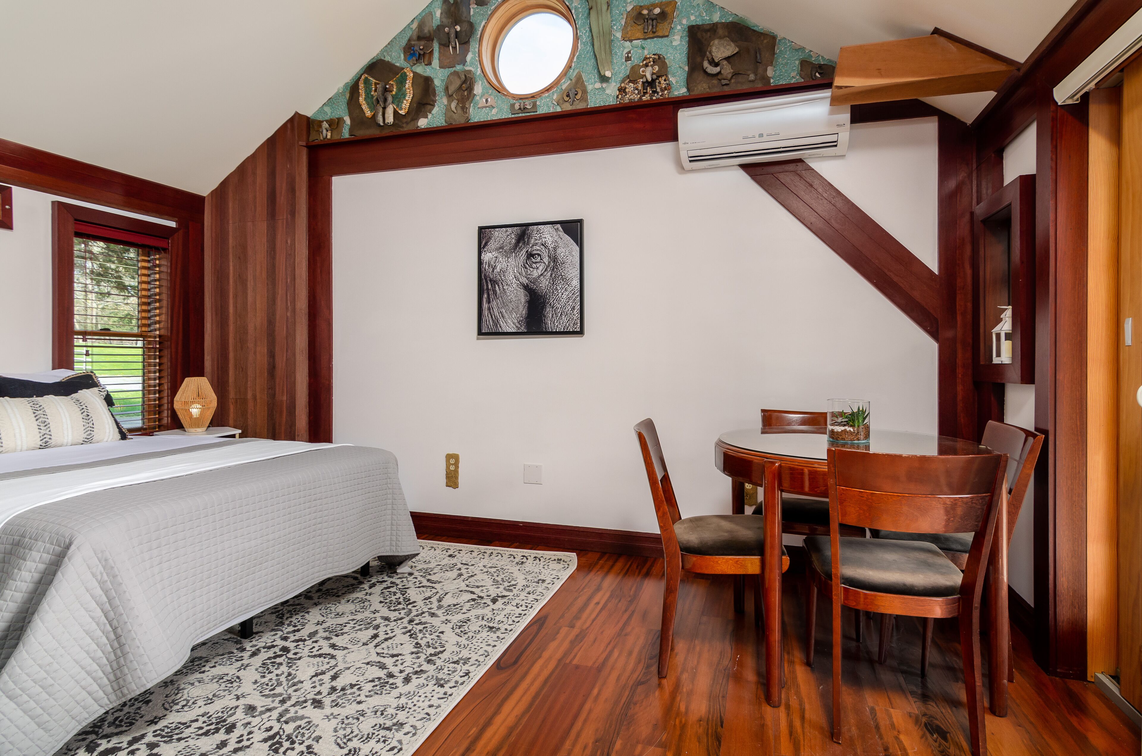 A bedroom with a bed, a desk, and a chair. There is a window with blinds, a wall-mounted air conditioner, and a framed picture on the wall.