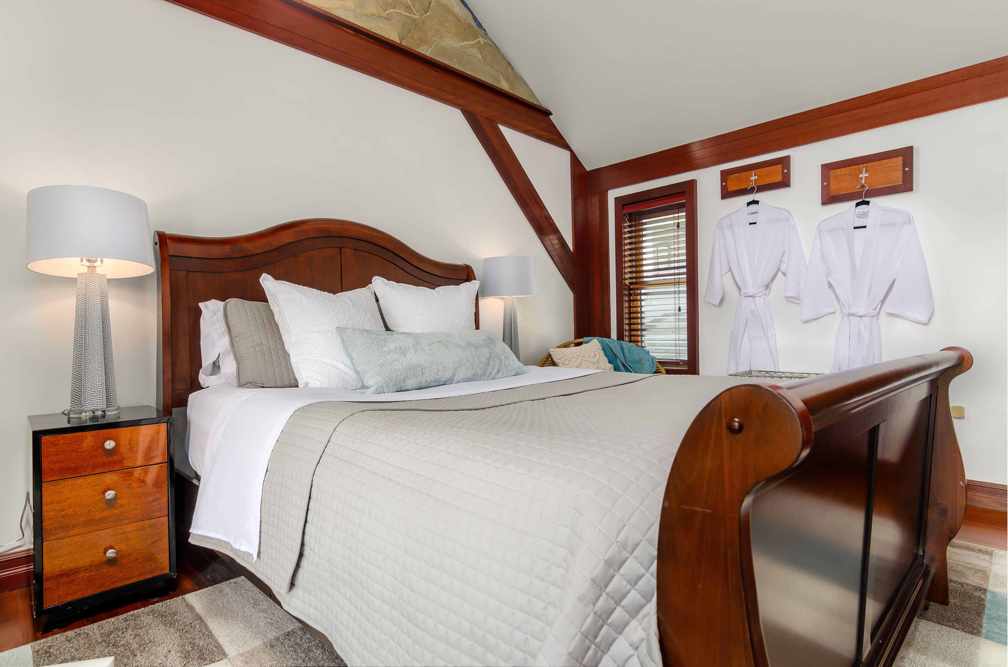 A neatly arranged bedroom with a wooden headboard, a bedside table with lamps, and two robes hanging on the wall.
