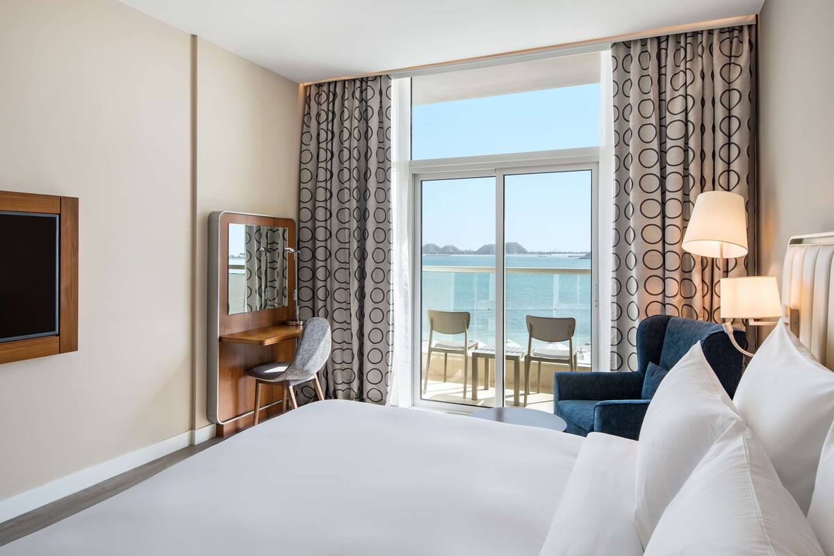 A hotel room with a large bed, a desk with a chair, a TV, a balcony with a view of the sea, and a lamp.