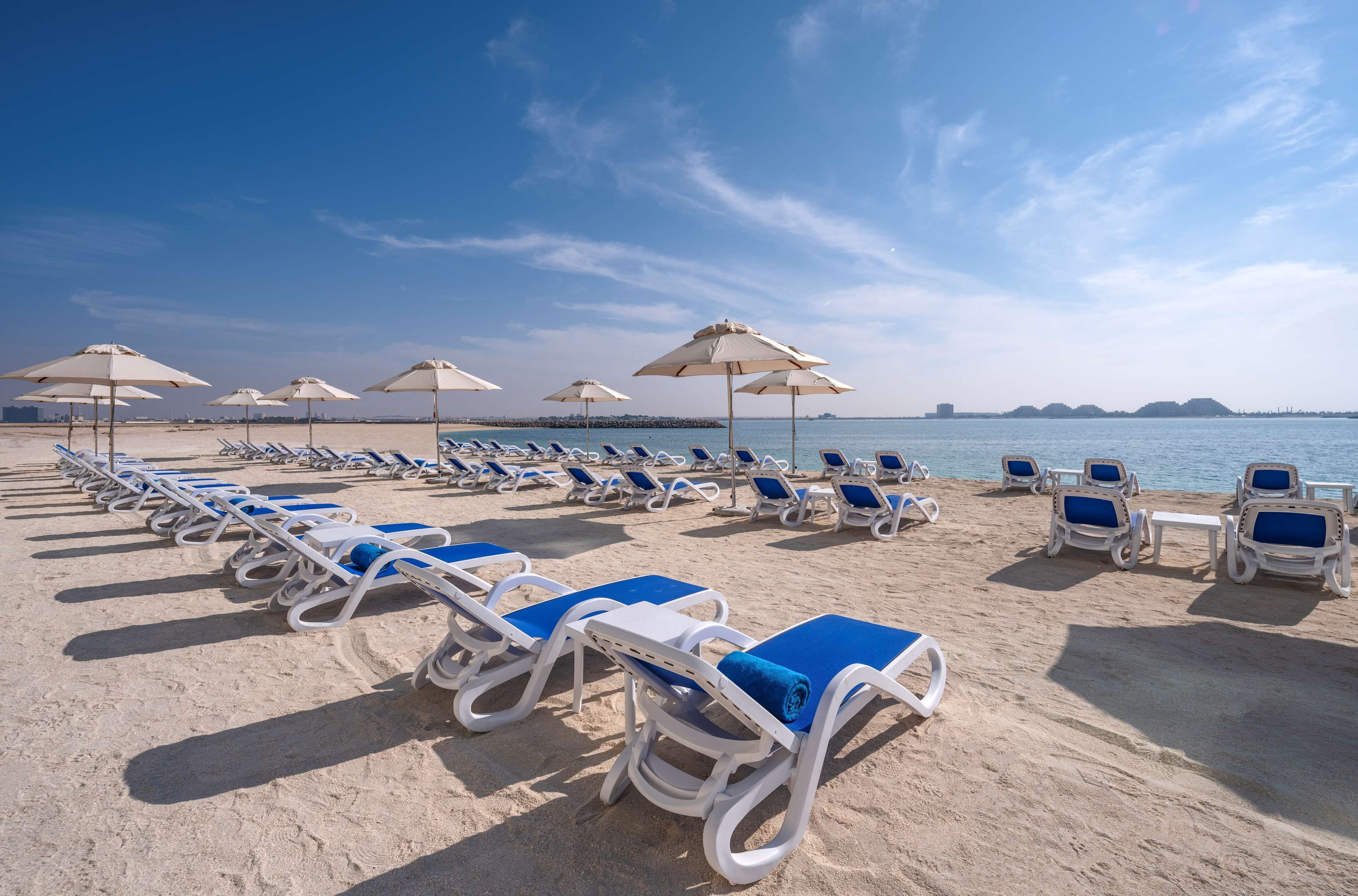 Une plage bordée de rangées de transats et de parasols, une plage de sable fin, et une mer calme en arrière-plan.