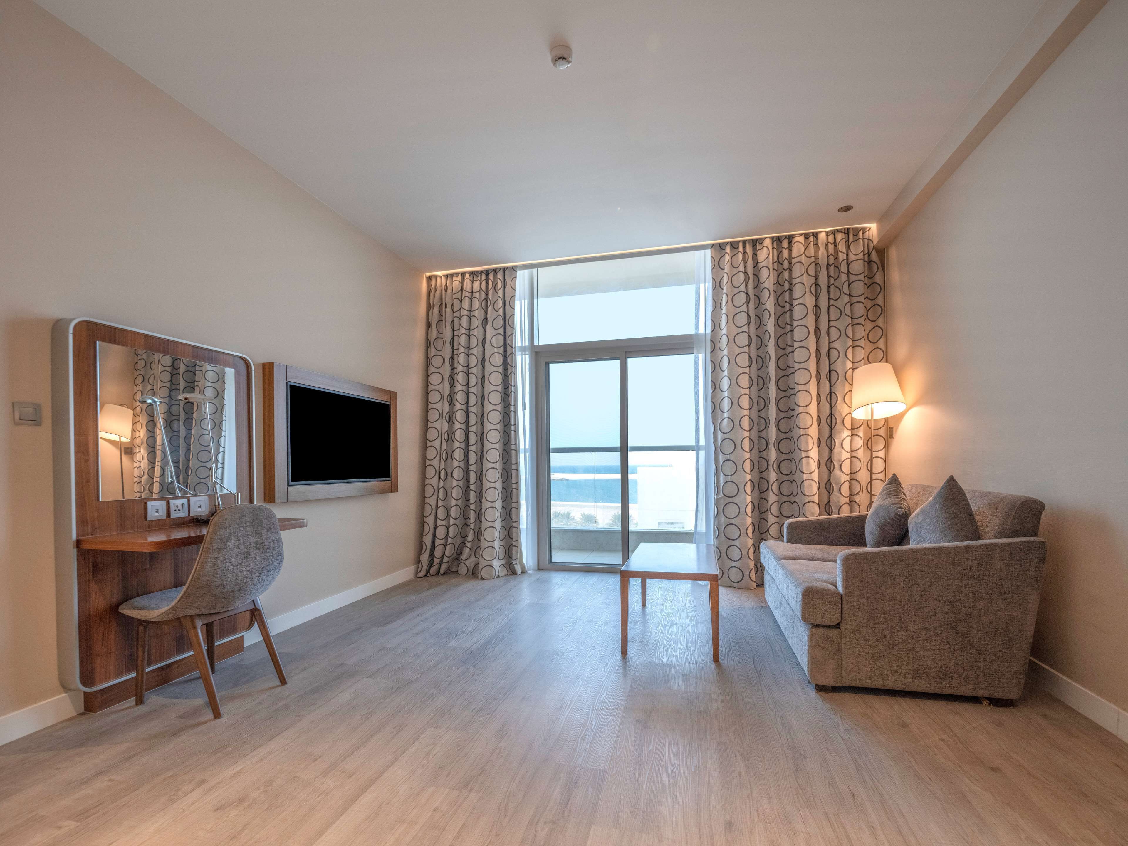 Een moderne hotelkamer met een houten bureau, een flatscreen televisie, een comfortabele bank en een groot raam met gordijnen.