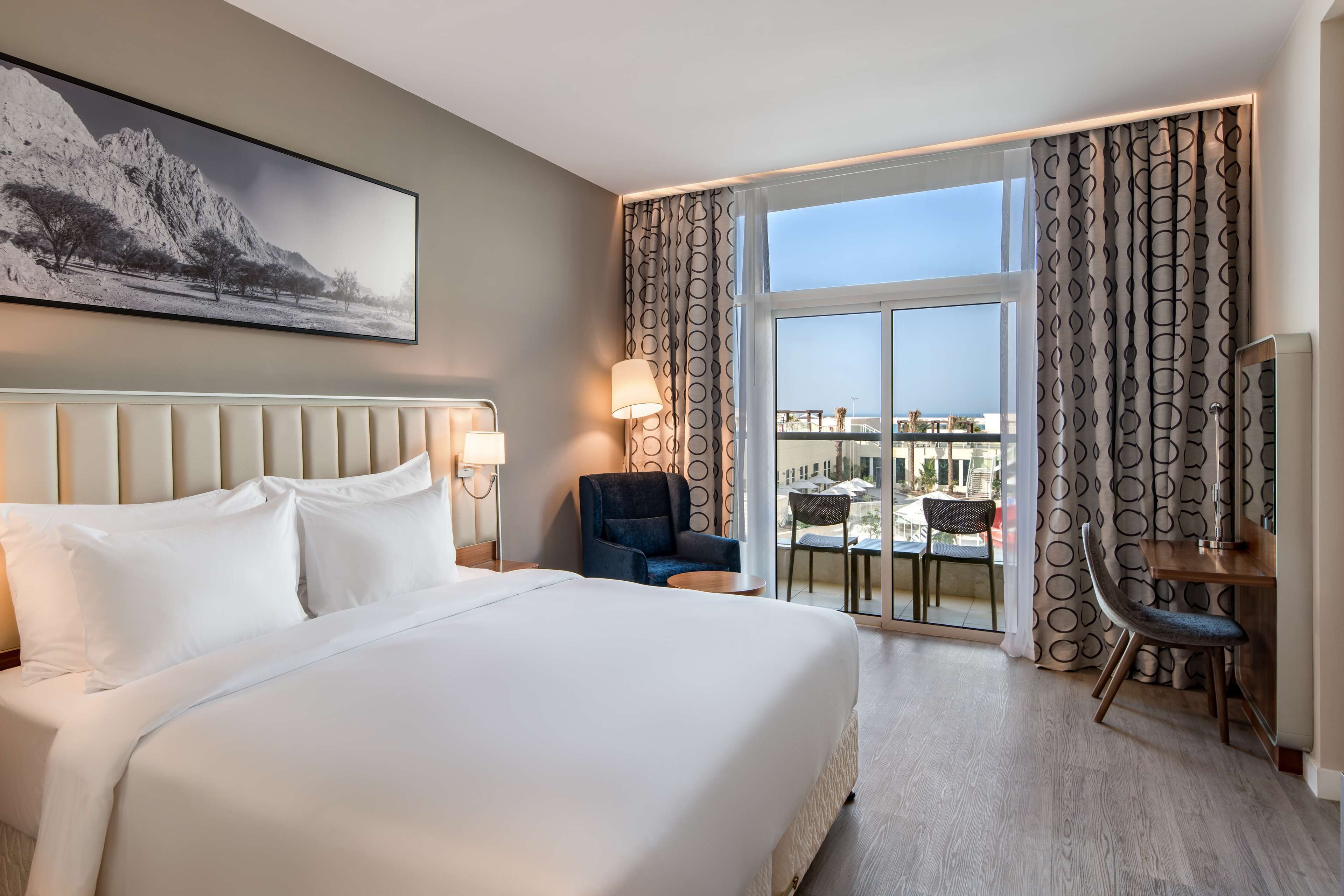 A hotel room with a large bed, a desk with a chair, a view of the city, and a balcony with a table and chairs.
