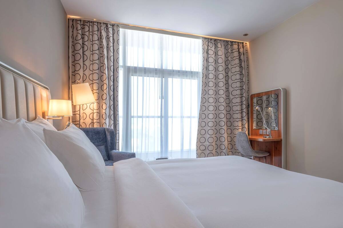 A hotel room with a large bed, a desk with a chair, a lamp, and a window with curtains.