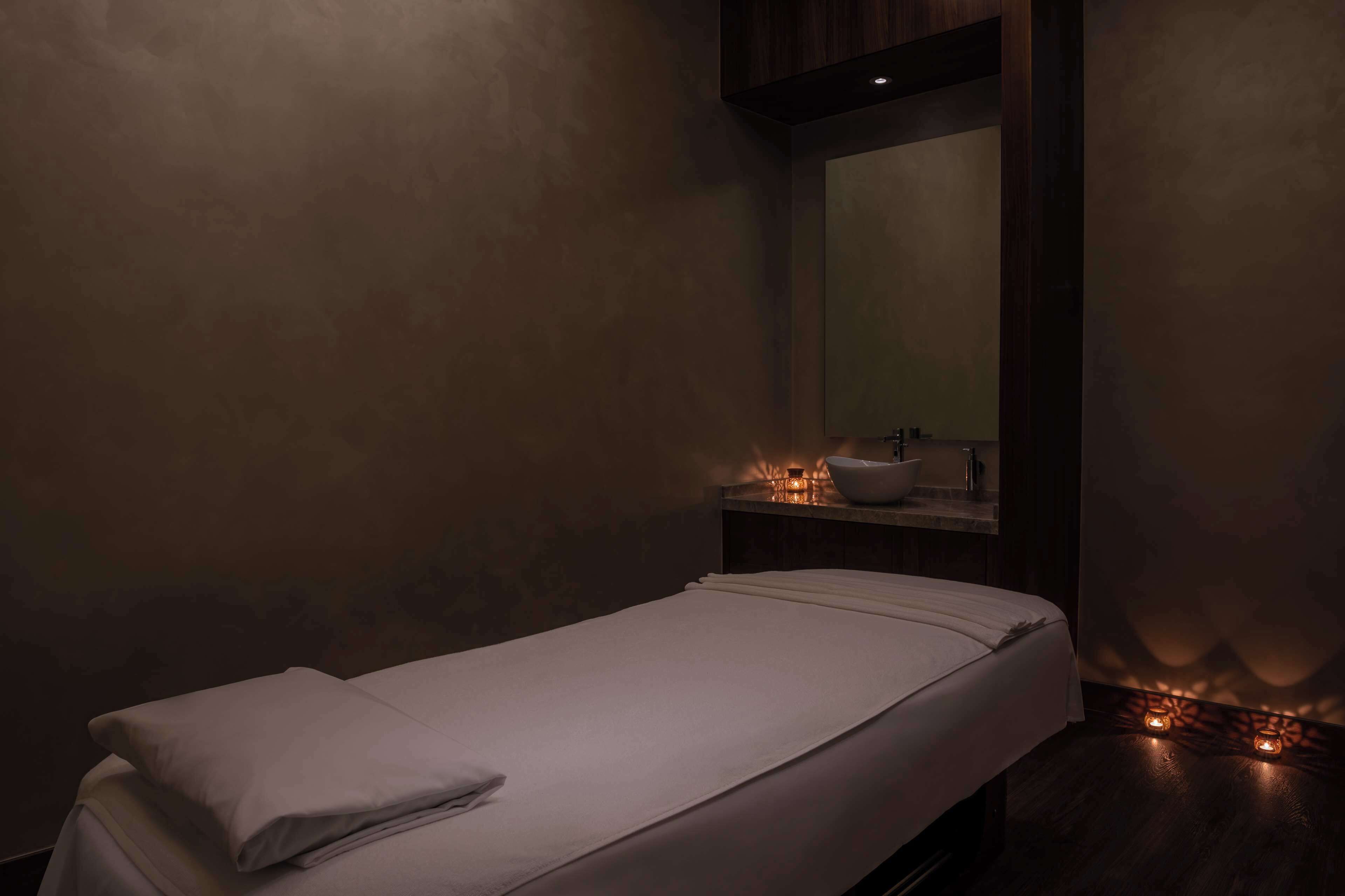 Una stanza poco illuminata con un letto singolo, un comodino in legno con uno specchio e una mensola a parete con candele.