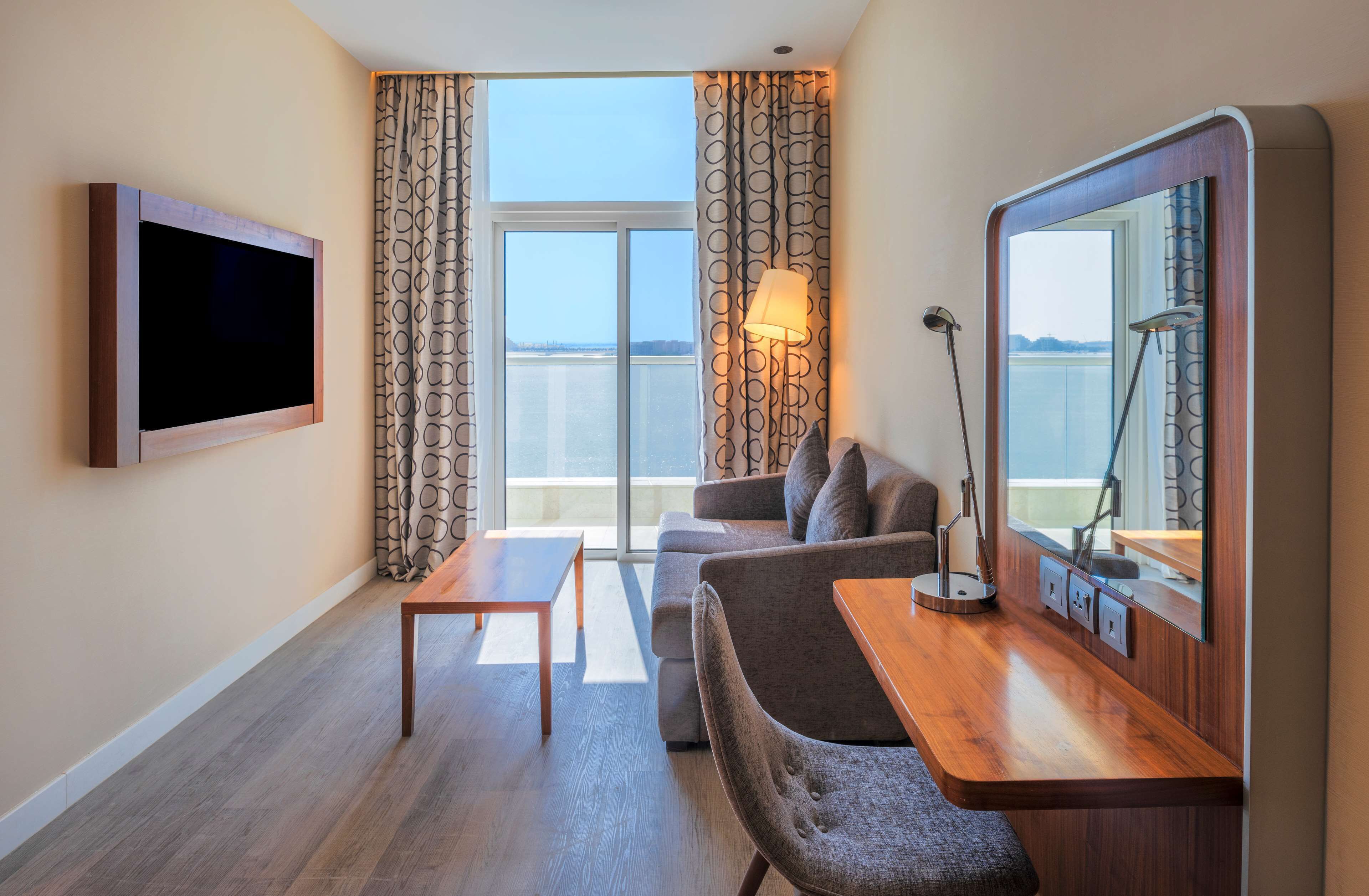 Een hotelkamer met een groot raam, een flatscreen televisie, een bank, een klein tafeltje en een spiegel.