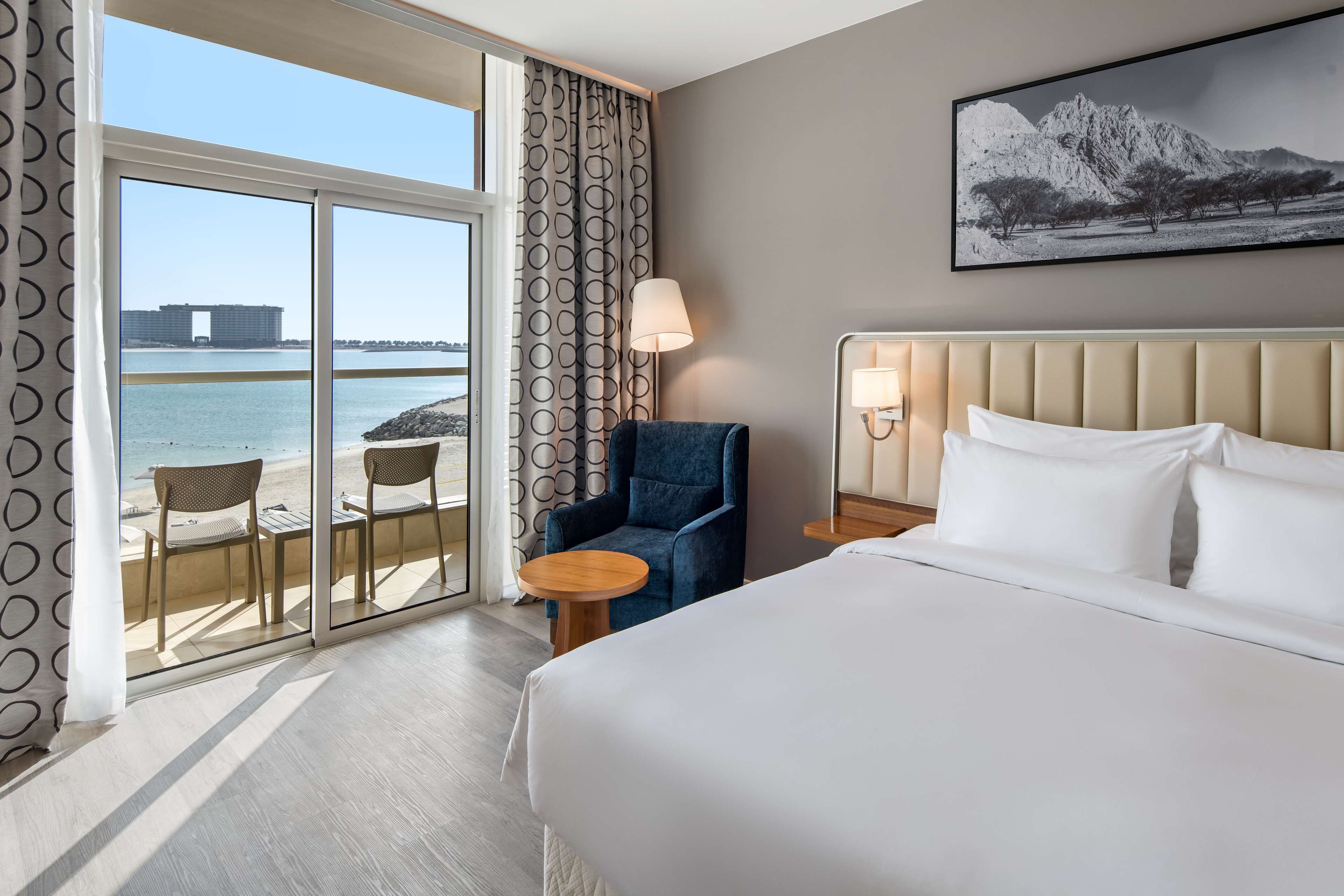 Une chambre d’hôtel avec un grand lit, une chaise, une petite table et une vue sur la mer, visible à travers une porte vitrée.