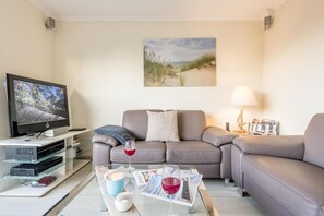 Uma sala de estar moderna com uma televisão de tela plana, um sofá cinza, uma mesa de centro com copos de vinho e uma pintura de uma praia.