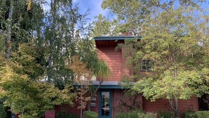 A two-story brick house with a front porch, surrounded by lush greenery and flowering plants.