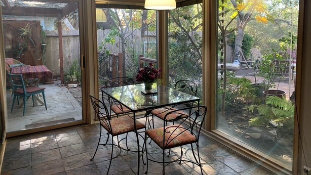 A dining area with a glass table and chairs, a hanging light fixture, and large windows offering a view of the garden.