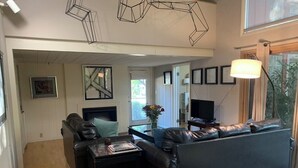 A modern living room with a black leather sofa, a coffee table, and a TV. There is a large window with curtains and a floor lamp.