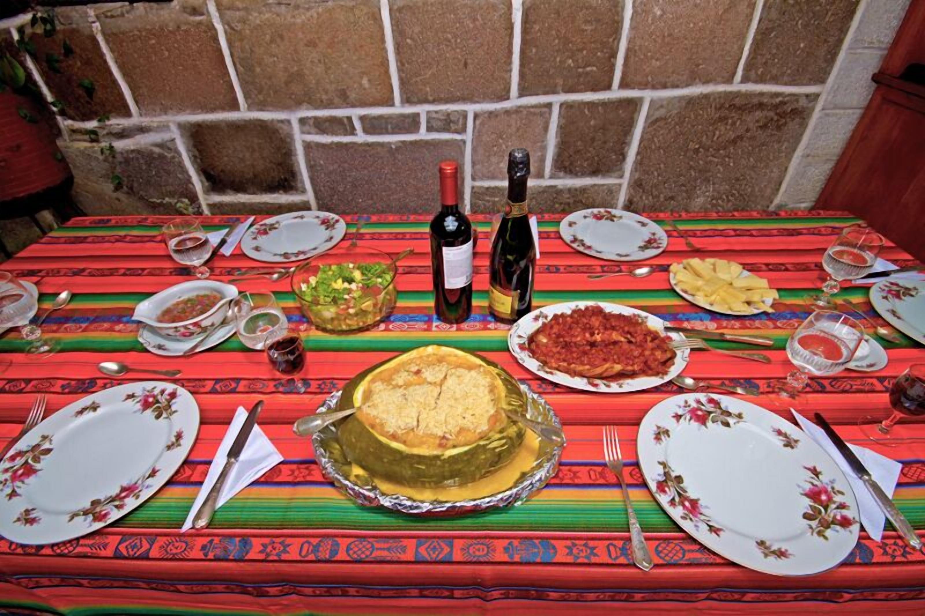Una mesa dispuesta con platos, cubiertos y una botella de vino, con un mantel colorido y una pared de piedra al fondo.