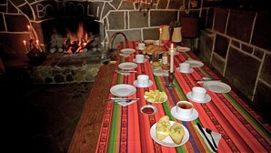 Een lange tafel gedekt voor een maaltijd, met kaarsen, borden en brood, voor een stenen openhaard met een 'BOMROLLI' bord.