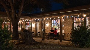An outdoor seating area with stone walls and string lights, surrounded by trees and a large tree trunk.