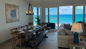 A dining area with a long table set for six, wooden chairs, and a bench. Large windows offer a view of the ocean.