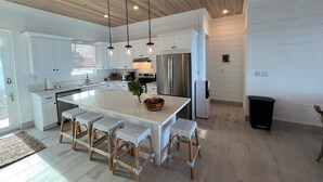 A modern kitchen with white cabinets, a large island with bar stools, stainless steel appliances, and a wooden ceiling.