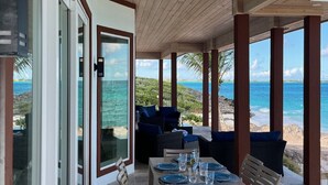 An outdoor dining area with a table set for two, overlooking a beach with clear blue water and rocky terrain.