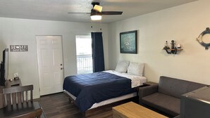Ein Schlafzimmer mit Bett, Schreibtisch, Stuhl, Deckenventilator, Fenster mit Vorhängen, einem Wandbild und einer kleinen Ablage mit Dekoration.