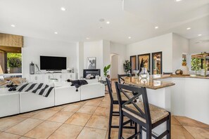 Una sala de estar moderna con un sofá blanco, una zona de comedor con una mesa y sillas de madera, un televisor de pantalla plana montado en la pared y una cocina con encimera y armarios.