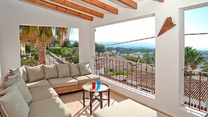 A balcony with a wicker sofa, a small round table, and a view of a residential area with palm trees and houses. - El Olivo by Interhome (Moraira)