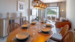 A dining area with a wooden table set for four, surrounded by wicker chairs. There is a large window providing a view of greenery outside. - Vacation home Melon by Interhome (La Herradura)