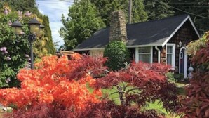 Une maison avec une cheminée en pierre, un porche et un jardin aux feuillages rouges.