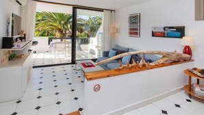 Een moderne woonkamer met een groot raam, een bank, een salontafel en een eethoek die zichtbaar is door de glazen deur.