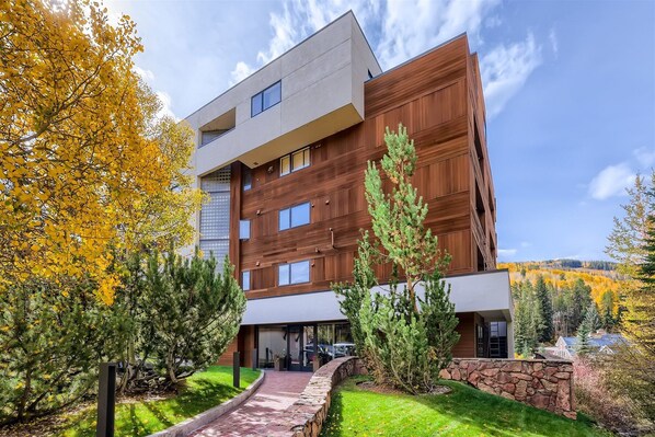 A modern multi-story building with a mix of wood paneling and concrete, surrounded by autumn foliage and a stone pathway.