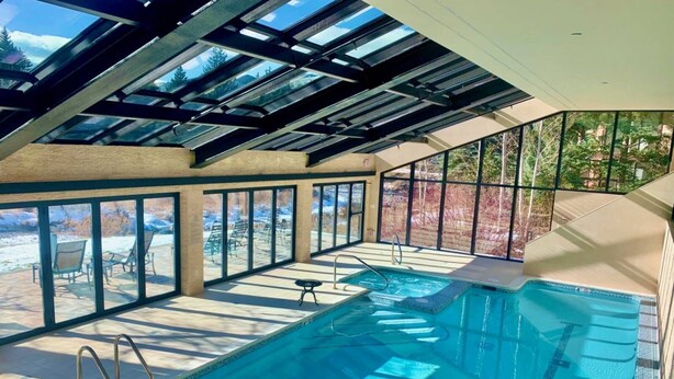 Una piscina interior con techo de cristal, vistas despejadas al exterior y una terraza soleada con sillas.