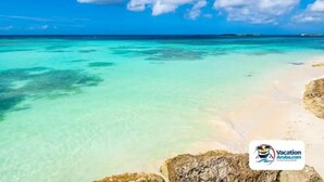Une plage aux eaux turquoise et au sable blanc, avec le logo de Vacation Aruba.com dans le coin inférieur droit.