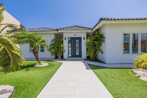 Une maison moderne avec un toit en tuiles, une plaque portant le numéro « 15A », et une pelouse bien entretenue agrémentée de palmiers.