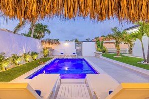 Une piscine dotée d’une terrasse blanche, entourée de palmiers et surmontée d’un toit de chaume.