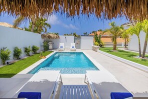 A rectangular swimming pool with white lounge chairs and a grill, surrounded by palm trees and a white wall.