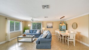 A living room with a blue sofa set, a dining table with chairs, a ceiling fan, and a large window with blinds. -  **GEM!**3 BR BEACH HOUSE w/ HEATED POOL, short ride to the beach!  (Naples)