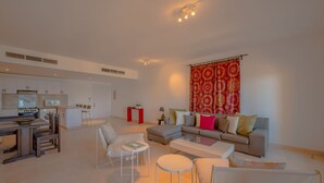 A modern living room with a sofa, coffee table, and chairs. There is a kitchen area with cabinets and appliances. A red curtain hangs in the background. - VESTA - Superior APT - 3BR - New Marina (El Gouna)
