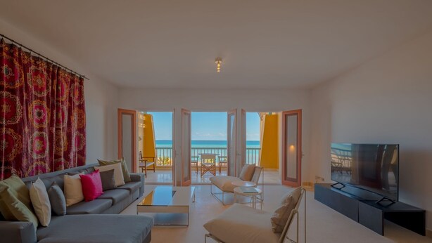 A spacious living room with a grey sofa, a white armchair, and a flat-screen TV on a black stand. A large window offers a view of the sea. - VESTA - Superior APT - 3BR - New Marina (El Gouna)