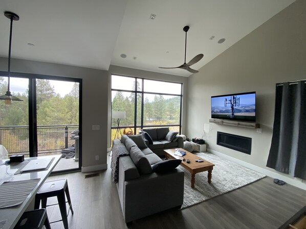 A modern living room with a sectional sofa, a coffee table, a flat-screen TV mounted on the wall, and a balcony with a view of trees.