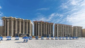 Un hotel frente a la playa con varios edificios altos, una playa arenosa con sombrillas azules y un cielo azul despejado.