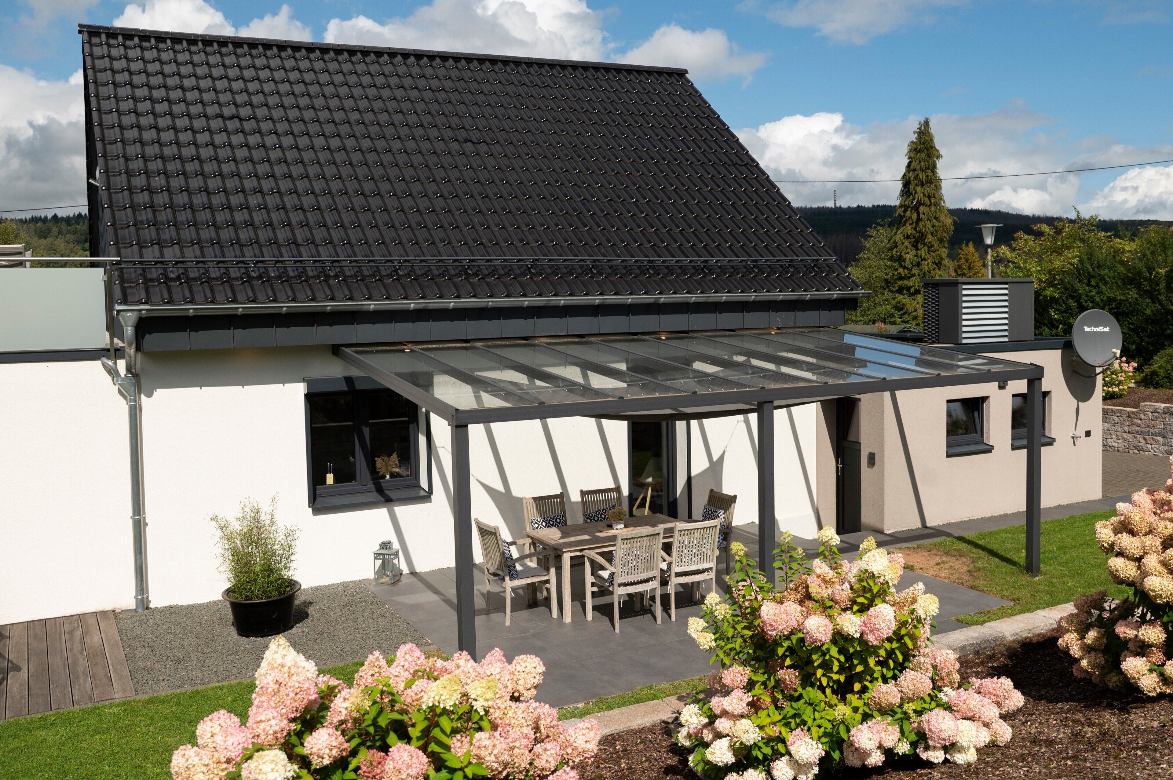A modern house with a covered patio, a black roof, and a garden with pink flowers.