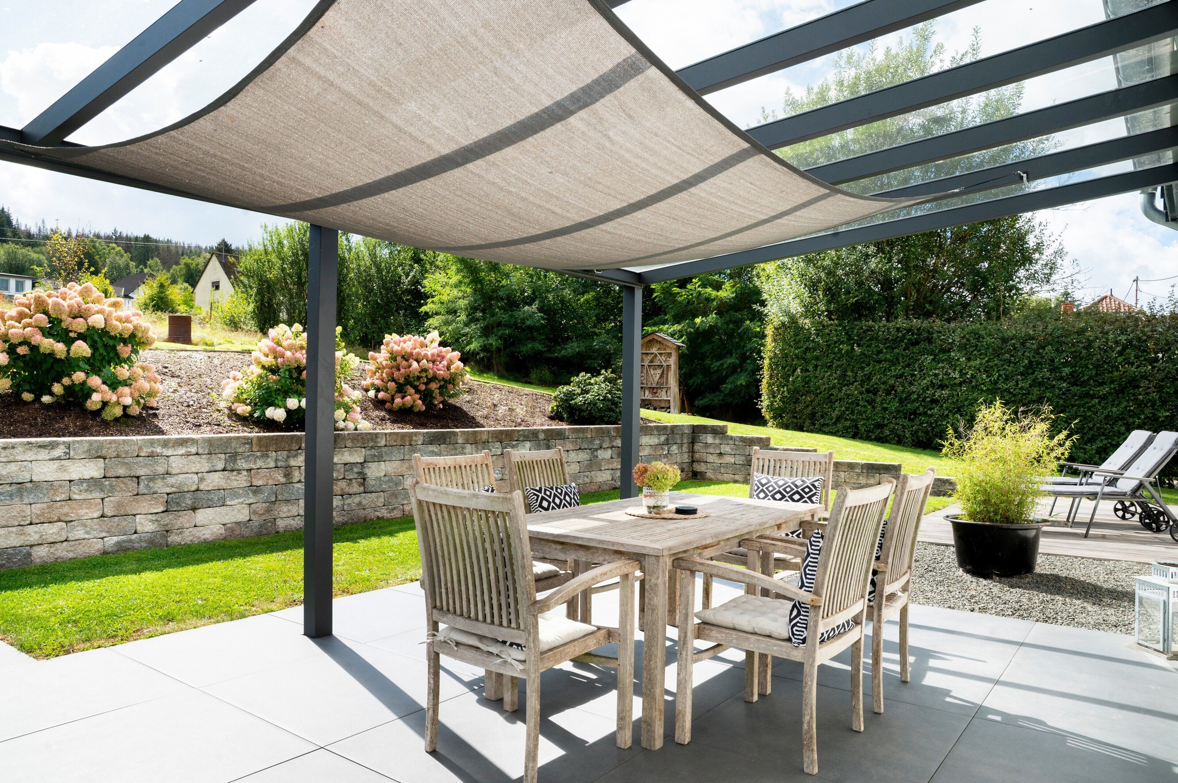 A covered patio with a table and chairs, a stone wall, and a garden with flowers and shrubs.