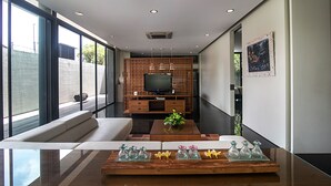Ein modernes Wohnzimmer mit einer Couch, einem Couchtisch mit Dekoration, einem Fernseher auf einem Holzschrank und großen Fenstern.