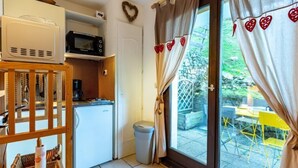 A compact kitchen with a microwave, a small refrigerator, and a coffee maker on a wooden stand. There is a trash bin next to the door, and a window with curtains decorated with heart shapes.