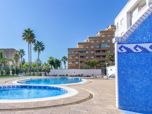 A swimming pool area with two pools, surrounded by palm trees and a building with a blue-tiled facade.