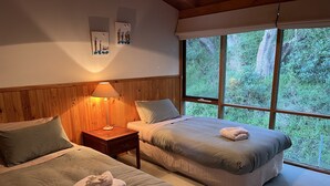 A room with two beds, a nightstand with a lamp, and a window with a view of greenery. - Perfect Waterfront Escape in Marlo | Balcony Views & Nature All Around (Marlo)