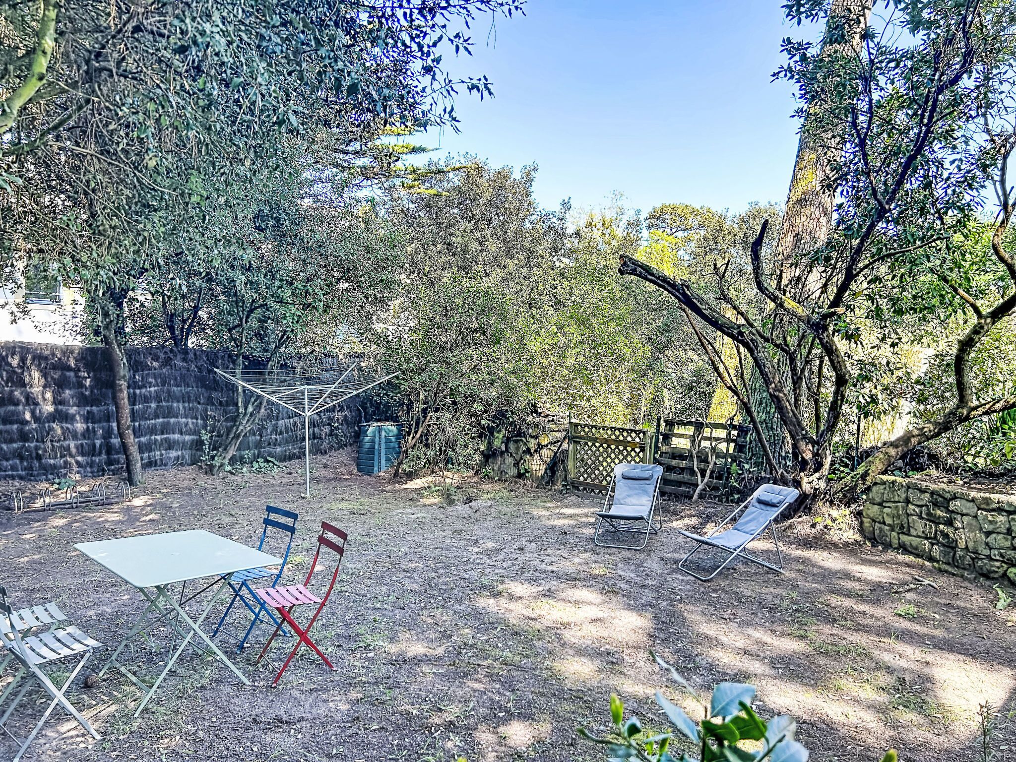 Un espace extérieur aménagé avec des tables et des chaises en métal, entouré d’arbres et d’un mur en pierre.