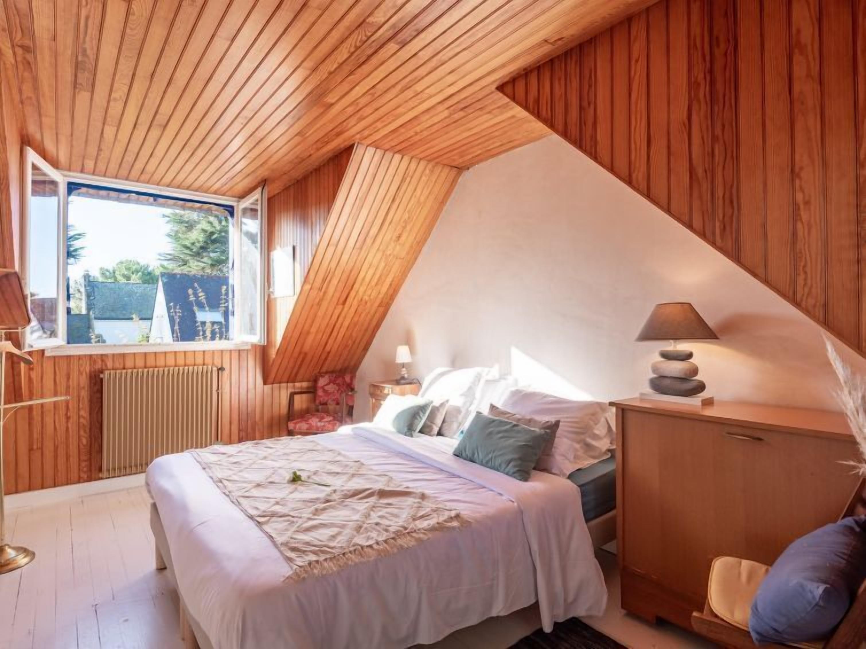 A bedroom with a sloped ceiling, wooden walls, a bed with white bedding, a bedside table with a lamp, and a view of the outside through a window.