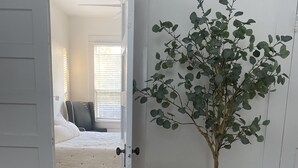 Une pièce avec un fauteuil gris, un coussin blanc, une plante en pot et une porte donnant sur une autre pièce où se trouvent un lit et des stores aux fenêtres.